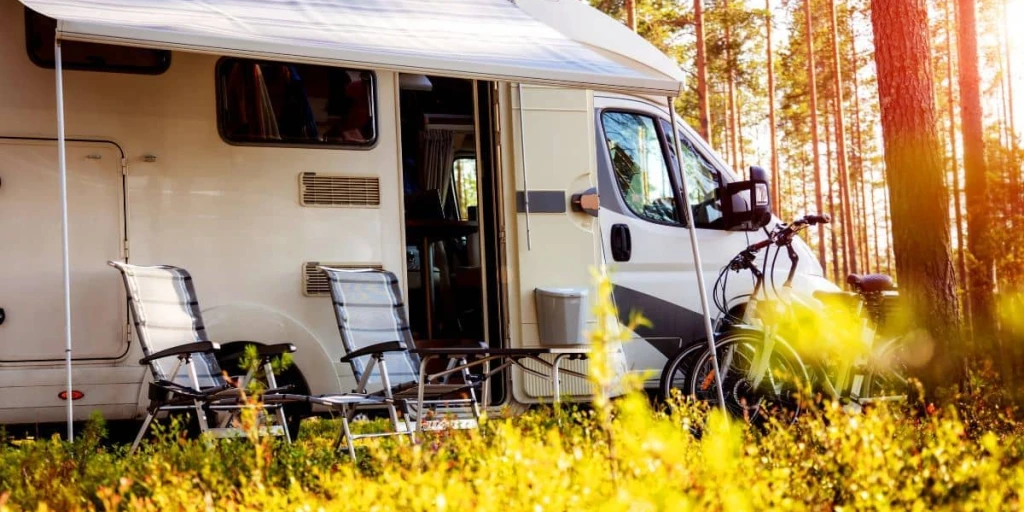 Campingwagen steht in einem Waldstück. Tür ist geöffnet und 2 Stühle und Fahrräder befinden sich davor. Die Sonne blitzt durch Nadelbäume und setzt die Kulisse in einem perfekten licht.